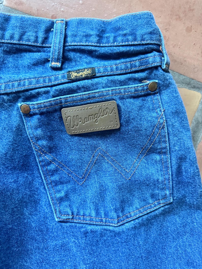 Puede incluir: Vaqueros de mezclilla azul medio con un parche rectangular de cuero marr&oacute;n en el bolsillo trasero que muestra el logotipo de Wrangler en cursiva. El bolsillo incluye remaches met&aacute;licos de color cobre y una costura decorativa en forma de W.