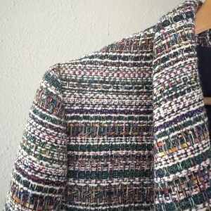 May include: Close-up of a multi-colored tweed blazer. The jacket features a woven pattern with threads of green, white, purple, and gold. The blazer is hanging on a wooden hanger.