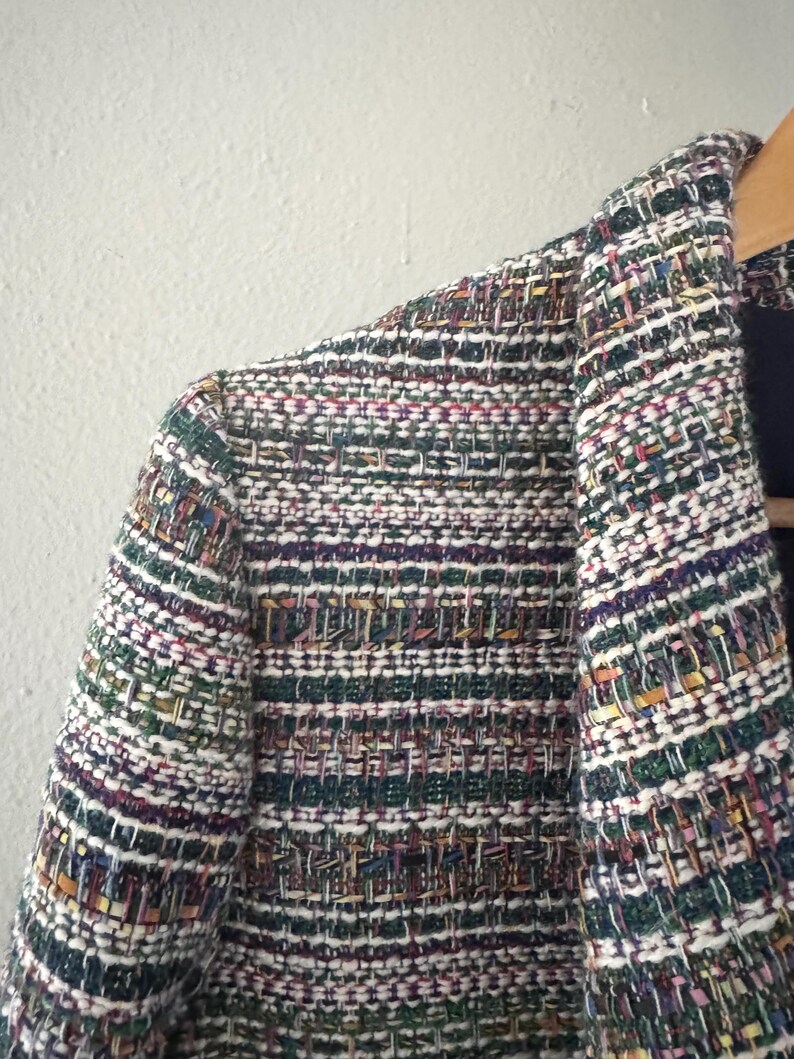 May include: Close-up of a multi-colored tweed blazer. The jacket features a woven pattern with threads of green, white, purple, and gold. The blazer is hanging on a wooden hanger.