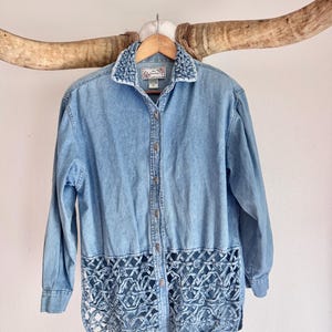May include: A light blue denim button-down shirt featuring a woven lattice design along the bottom hem and collar. The shirt has long sleeves, a standard collar, and a front button closure. The fabric has a faded wash appearance throughout.