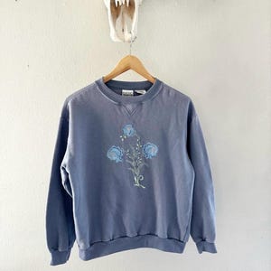 May include: A light blue sweatshirt with a floral design featuring blue flowers and green leaves. The sweatshirt has a crew neck and long sleeves.