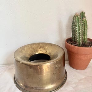 May include: A vintage brass ashtray with a wide, circular opening and a slightly flared base. The ashtray has a tarnished, aged appearance, suggesting it is an antique. A small potted cactus sits nearby.