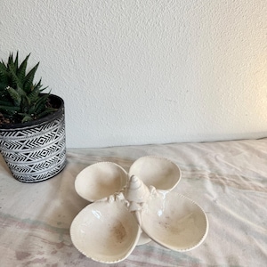 May include: Off-white ceramic serving dish with four petal-shaped compartments. The dish has a textured surface and a central, cone-shaped accent. A potted succulent plant is in the background.