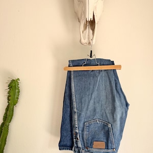 K&ouml;nnte beinhalten: Blaue Jeans aus Denim, die an einem Holzb&uuml;gel h&auml;ngen. Die Jeans haben ein klassisches F&uuml;nf-Taschen-Design und einen braunen Lederaufn&auml;her auf der Ges&auml;&szlig;tasche. &Uuml;ber der Jeans h&auml;ngt ein Kuhsch&auml;del an einer wei&szlig;en Wand.