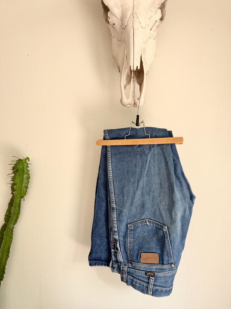 K&ouml;nnte beinhalten: Blaue Jeans aus Denim, die an einem Holzb&uuml;gel h&auml;ngen. Die Jeans haben ein klassisches F&uuml;nf-Taschen-Design und einen braunen Lederaufn&auml;her auf der Ges&auml;&szlig;tasche. &Uuml;ber der Jeans h&auml;ngt ein Kuhsch&auml;del an einer wei&szlig;en Wand.