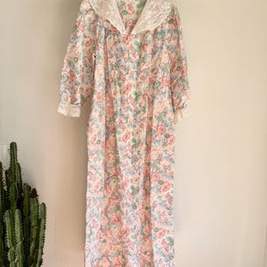 May include: A vintage floral print nightgown with a lace collar and cuffs. The gown features a pattern of pink and red roses with green leaves on a cream background. The long-sleeved gown is hanging on a wooden hanger.