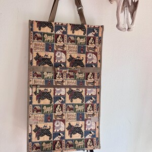 May include: A rectangular rolling shopping bag featuring a woven tapestry pattern of various dog breeds, dog houses, and the text Faithful Companion. The bag has brown fabric trim, two adjustable brown straps, and a horizontal zippered front pocket.