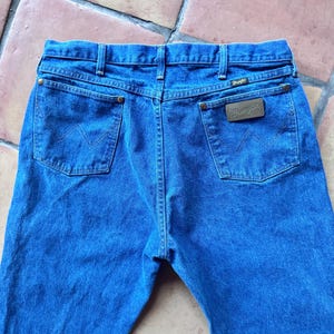 Puede incluir: Unos vaqueros de mezclilla azul con un dise&ntilde;o cl&aacute;sico de cinco bolsillos, cintura con trabillas para cintur&oacute;n y un parche de cuero marr&oacute;n en el bolsillo trasero derecho con el nombre de la marca Wrangler. Los bolsillos traseros tienen costuras en W.