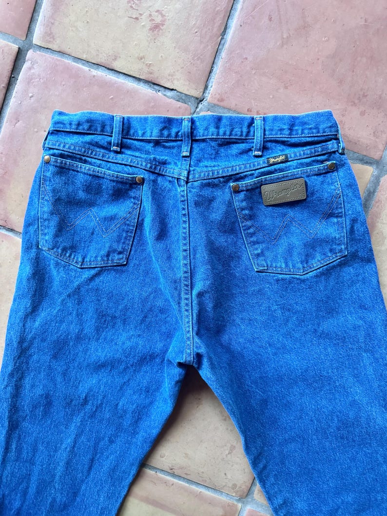 Puede incluir: Unos vaqueros de mezclilla azul con un dise&ntilde;o cl&aacute;sico de cinco bolsillos, cintura con trabillas para cintur&oacute;n y un parche de cuero marr&oacute;n en el bolsillo trasero derecho con el nombre de la marca Wrangler. Los bolsillos traseros tienen costuras en W.