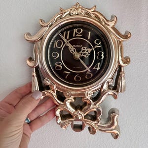 May include: A gold-colored quartz wall clock with a black face and ornate detailing. The clock face features gold numerals and hands, with the brand name "MANIX" visible. The clock is held by a hand.