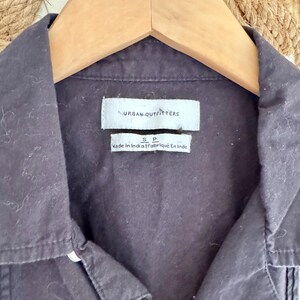 May include: Close-up of a dark gray button-down shirt hanging on a wooden hanger. The shirt has a collar and a small tag that reads "URBAN OUTFITTERS". Below the tag is another tag that reads "Made In India | Fabriqu&eacute; En Inde".