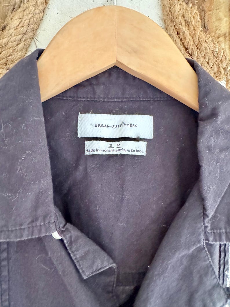 May include: Close-up of a dark gray button-down shirt hanging on a wooden hanger. The shirt has a collar and a small tag that reads "URBAN OUTFITTERS". Below the tag is another tag that reads "Made In India | Fabriqu&eacute; En Inde".