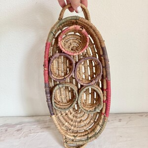 May include: Woven oval wall basket with a handle. The basket features a natural beige color with accents of pink, purple, and green. It has several circular openings and is made of natural fibers.