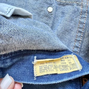 Puede incluir: Primer plano de unos vaqueros azules con una etiqueta visible. La etiqueta indica una talla de cintura de 81 cm y una longitud de 84 cm. Los vaqueros tienen un bot&oacute;n y costuras visibles.
