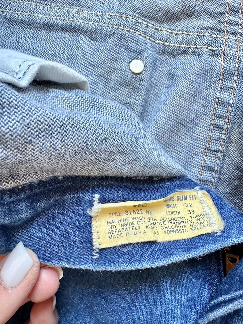 Puede incluir: Primer plano de unos vaqueros azules con una etiqueta visible. La etiqueta indica una talla de cintura de 81 cm y una longitud de 84 cm. Los vaqueros tienen un bot&oacute;n y costuras visibles.