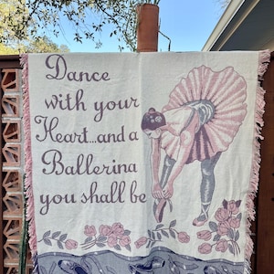 May include: Cream-colored throw blanket with a ballerina design. The blanket features a ballerina in a pink tutu, the text "Dance with your Heart...and a Ballerina you shall be", and a floral border. The edges are fringed.