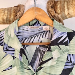Peut inclure: Chemise &agrave; imprim&eacute; feuilles tropicales vertes, noires et bleues, suspendue &agrave; un cintre en bois. L'&eacute;tiquette indique la marque "Tommy Bahama". La chemise est boutonn&eacute;e.