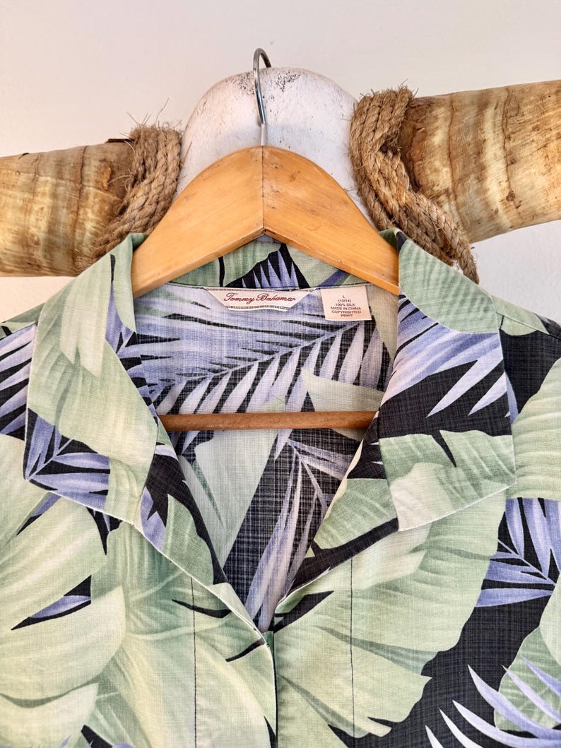 Peut inclure: Chemise &agrave; imprim&eacute; feuilles tropicales vertes, noires et bleues, suspendue &agrave; un cintre en bois. L'&eacute;tiquette indique la marque "Tommy Bahama". La chemise est boutonn&eacute;e.