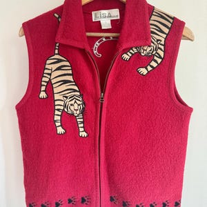 May include: A red, sleeveless zip-up vest with embroidered tiger designs. The vest features two tigers, one on each side, and paw prints along the bottom hem. The vest is hanging on a wooden hanger.