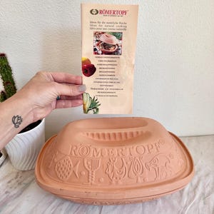 May include: A terracotta Romertopf clay baker with a lid featuring embossed fish, goblet, rooster, and pig motifs. The lid is inscribed with the brand name. An accompanying informational pamphlet displays the brand logo and cooking imagery.