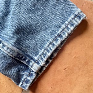 May include: Close-up of a section of light blue denim jeans. The hem is cuffed, revealing the raw edge of the fabric. The denim has a faded, worn appearance, with visible stitching along the seams.