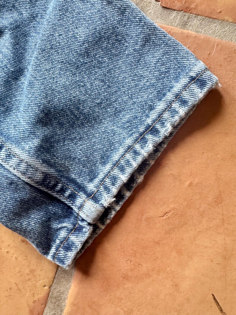 May include: Close-up of a section of light blue denim jeans. The hem is cuffed, revealing the raw edge of the fabric. The denim has a faded, worn appearance, with visible stitching along the seams.
