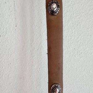 May include: Close-up of a brown leather belt with two silver oval decorative studs. The studs feature a detailed floral design around the edges. The belt is against a white textured background.