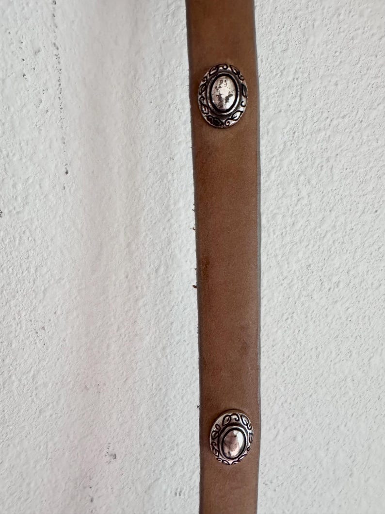 May include: Close-up of a brown leather belt with two silver oval decorative studs. The studs feature a detailed floral design around the edges. The belt is against a white textured background.