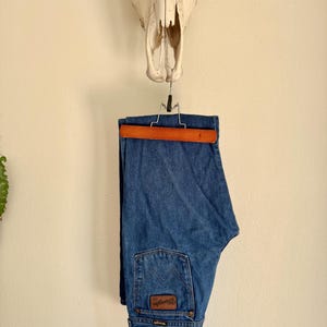 Puede incluir: Vaqueros azules de mezclilla colgados de una percha de madera. Los vaqueros tienen un parche de cuero marr&oacute;n en el bolsillo trasero con el logotipo de Wrangler. Un cr&aacute;neo de vaca est&aacute; montado encima de los vaqueros en una pared color crema.