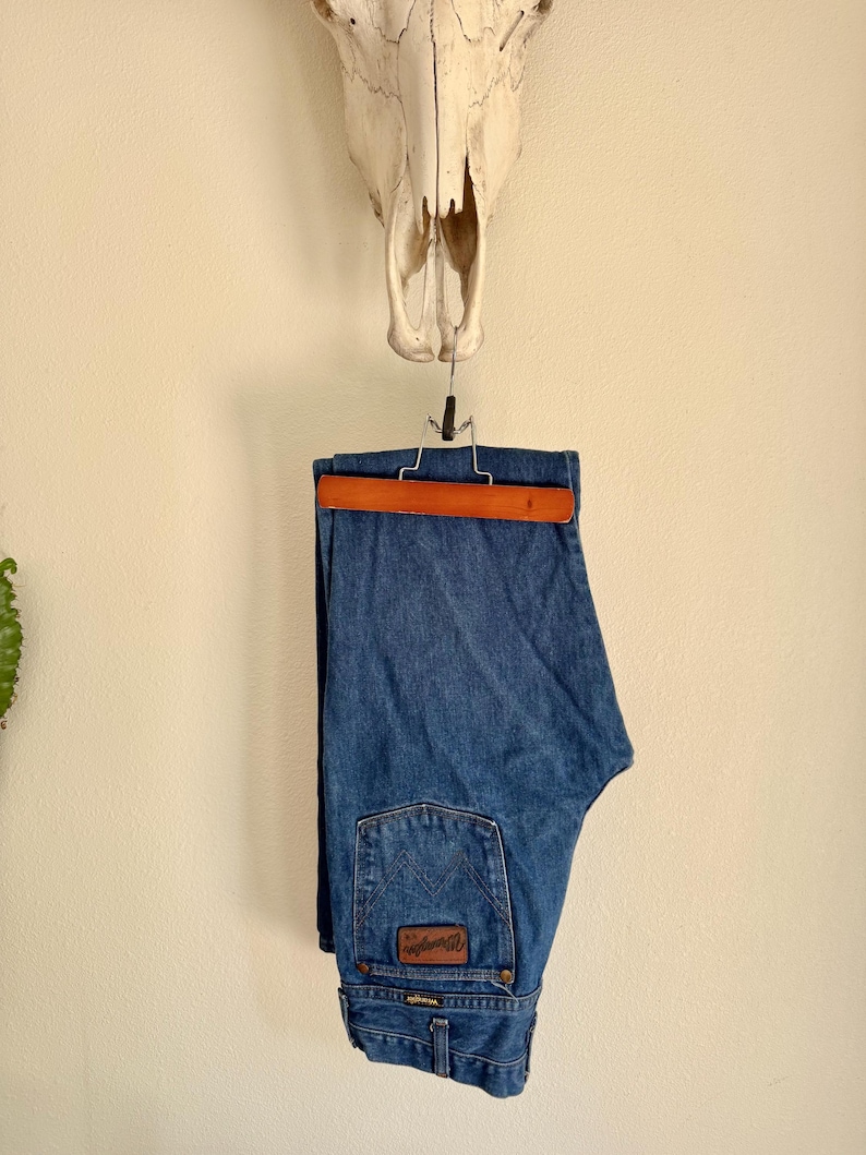 Puede incluir: Vaqueros azules de mezclilla colgados de una percha de madera. Los vaqueros tienen un parche de cuero marr&oacute;n en el bolsillo trasero con el logotipo de Wrangler. Un cr&aacute;neo de vaca est&aacute; montado encima de los vaqueros en una pared color crema.