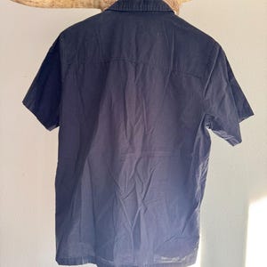 May include: A navy blue short-sleeved shirt hanging on a wooden hanger. The shirt has a button-down collar and a classic design. The back of the shirt is visible, showing the seams and the texture of the fabric. The shirt is hanging against a neutral background.