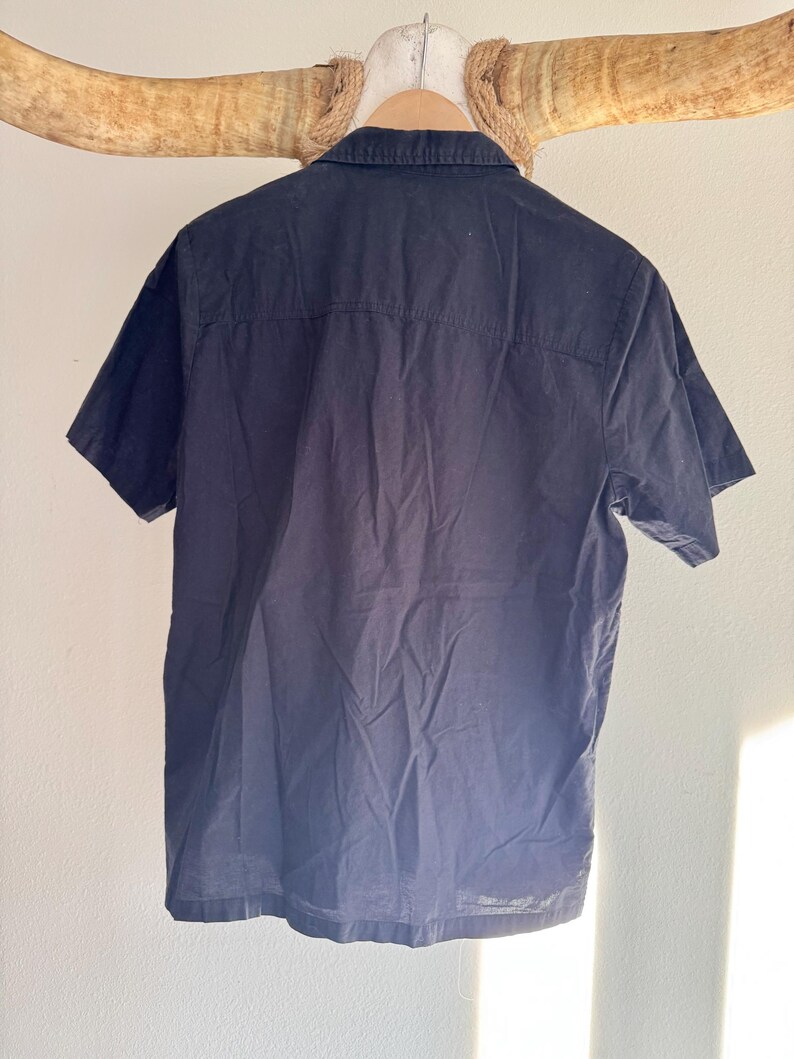 May include: A navy blue short-sleeved shirt hanging on a wooden hanger. The shirt has a button-down collar and a classic design. The back of the shirt is visible, showing the seams and the texture of the fabric. The shirt is hanging against a neutral background.