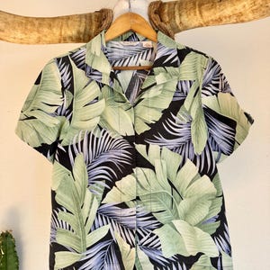Peut inclure: Chemise &agrave; manches courtes &agrave; boutons avec un motif de feuilles tropicales. La chemise pr&eacute;sente de grandes feuilles vertes et de plus petites frondes bleu-gris sur un fond noir. La chemise est suspendue &agrave; un cintre en bois.