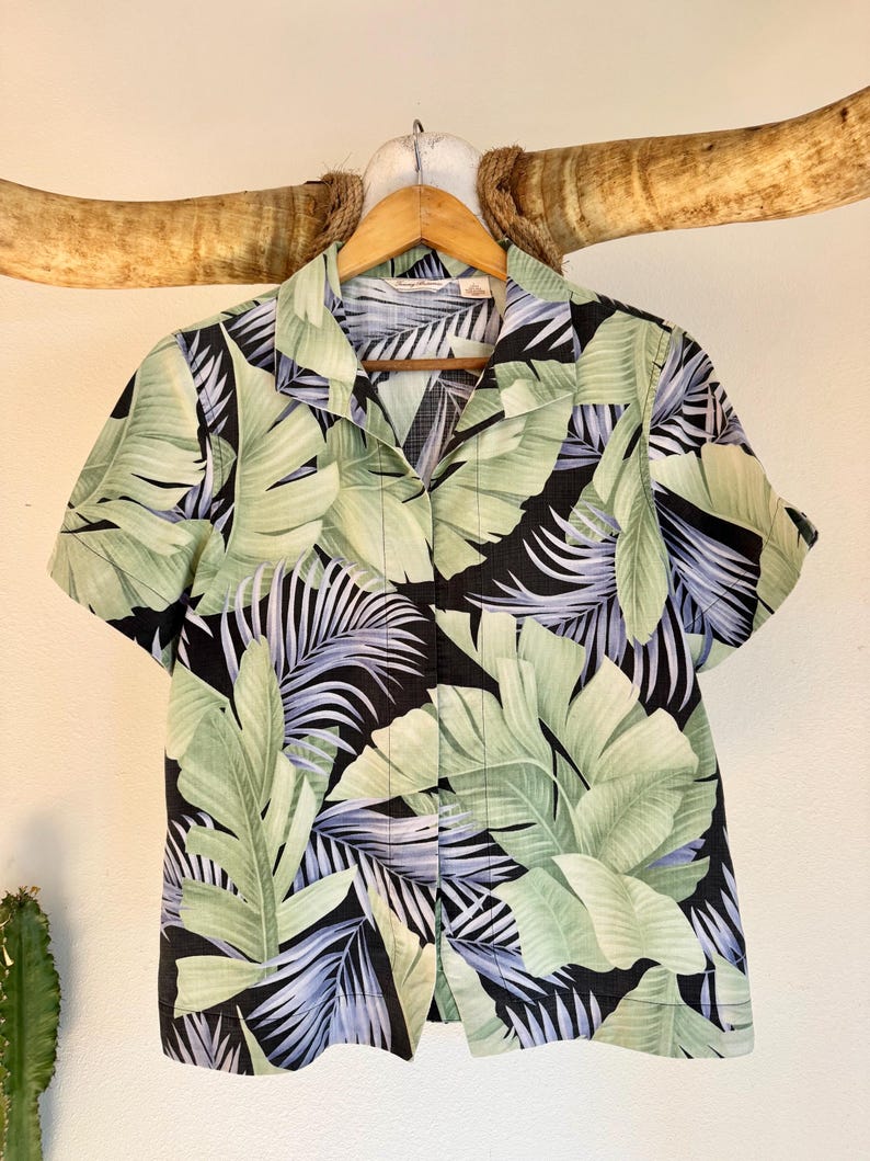 Peut inclure: Chemise &agrave; manches courtes &agrave; boutons avec un motif de feuilles tropicales. La chemise pr&eacute;sente de grandes feuilles vertes et de plus petites frondes bleu-gris sur un fond noir. La chemise est suspendue &agrave; un cintre en bois.