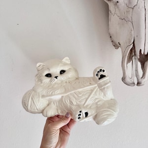 May include: A cream-colored ceramic cat figurine with a ball of yarn. The cat has black eyes and a detailed fur texture. The figurine is held up against a white wall, with a skull in the background.