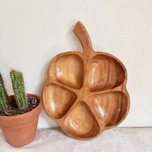 May include: A wooden serving tray with five compartments and a handle. The tray is shaped like a flower or a stylized fruit, with a natural wood grain finish. A small potted cactus is visible on the left side of the image.