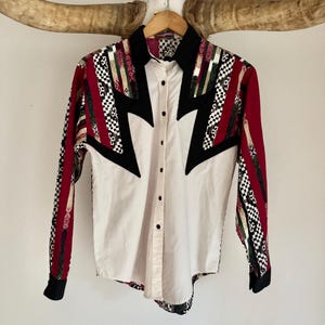 May include: A long-sleeved button-down shirt with a white front and patterned sleeves. The sleeves feature red, black, green, and white geometric designs. The shirt has a black collar and accents, and is hanging on a wooden hanger.