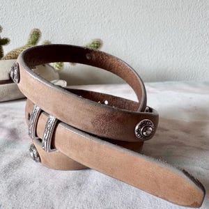 May include: A tan leather belt with silver metal studs and buckle detailing. The belt has a worn, vintage look. The leather belt is coiled, showing its design. The belt is made of leather.