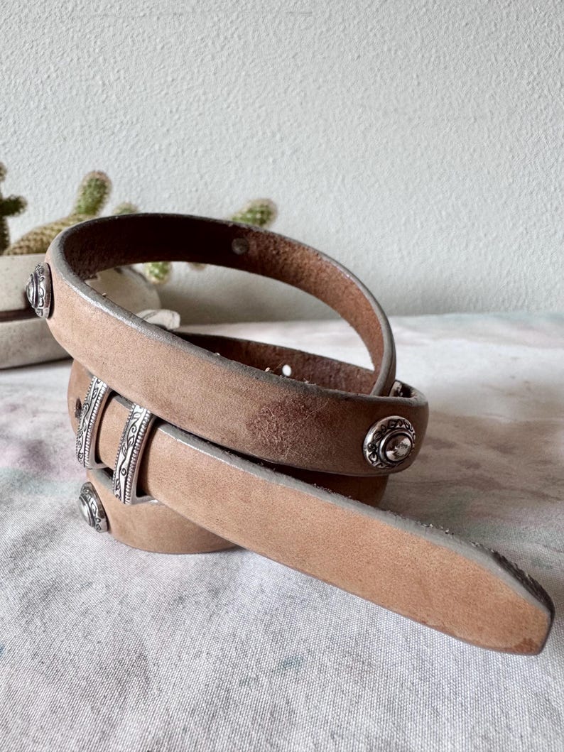 May include: A tan leather belt with silver metal studs and buckle detailing. The belt has a worn, vintage look. The leather belt is coiled, showing its design. The belt is made of leather.