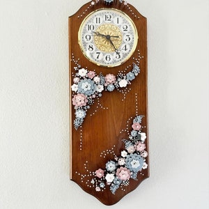 May include: A wooden clock with a white face and black numbers. The clock is decorated with pink and blue flowers made of fabric. The clock is hanging on a white wall.