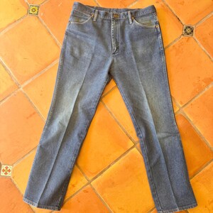 May include: Light blue denim jeans with a classic five-pocket design. The jeans are laid flat on a terracotta tile floor. The jeans have a button and zipper closure and belt loops.