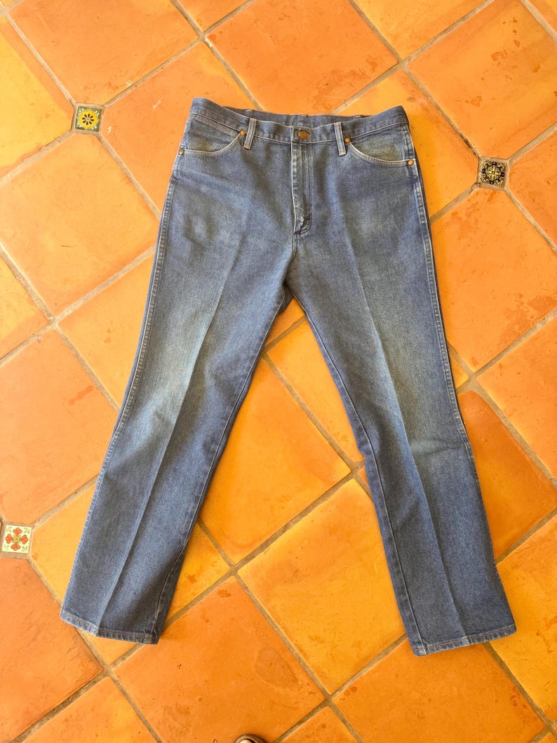 May include: Light blue denim jeans with a classic five-pocket design. The jeans are laid flat on a terracotta tile floor. The jeans have a button and zipper closure and belt loops.