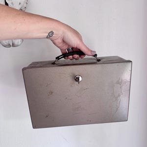 May include: A rectangular metal cash box with a weathered taupe finish, featuring a black plastic carrying handle on the top and a central silver-toned key lock mechanism. The surface shows visible signs of wear, including scratches and minor rust.