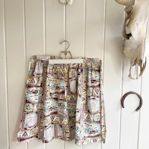May include: A brown and white patterned apron with a green, blue, and yellow design. The apron is hanging on a hanger against a white wood panel wall.