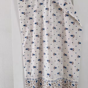May include: White fabric with a repeating pattern of blue teardrop shapes and brown diamond shapes. The bottom border features a design of blue teardrops and brown leaves. The fabric is hanging from a wooden hanger.