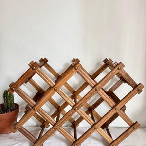 May include: A wooden wine rack with a crisscross design. The rack is made of light brown wood and features brass-colored rivets at each joint. The rack is designed to hold multiple bottles of wine and has a collapsible design.