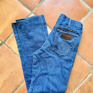 Puede incluir: Vaqueros Wrangler de mezclilla azul con un parche de cuero marr&oacute;n en el bolsillo trasero. Los vaqueros est&aacute;n colocados sobre un suelo de baldosas de terracota. El logotipo de Wrangler es visible en el bolsillo trasero.