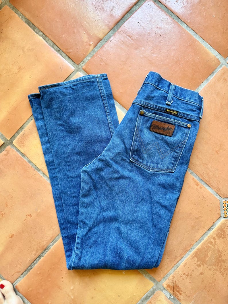 Puede incluir: Vaqueros Wrangler de mezclilla azul con un parche de cuero marr&oacute;n en el bolsillo trasero. Los vaqueros est&aacute;n colocados sobre un suelo de baldosas de terracota. El logotipo de Wrangler es visible en el bolsillo trasero.