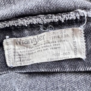 Op de afbeelding: Close-up van grijze denim stof met een wit Wrangler label. Het label bevat wasinstructies en materiaalinformatie, 100% katoen. Er staat ook een productcode op.
