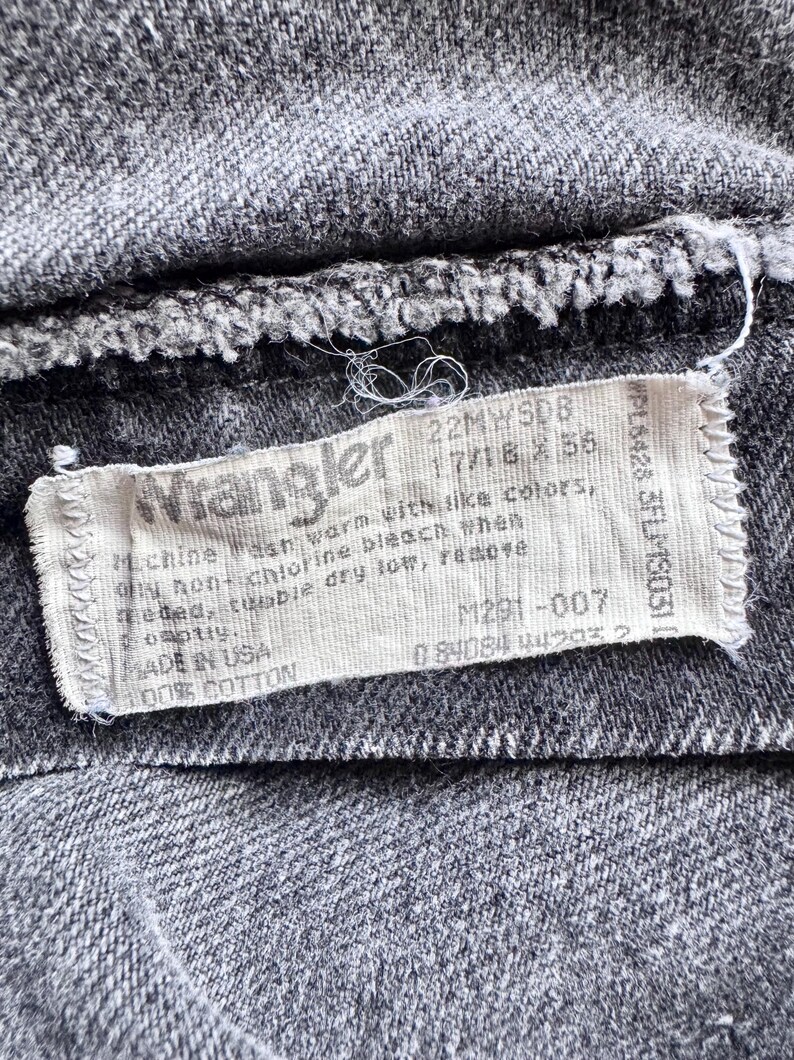 Op de afbeelding: Close-up van grijze denim stof met een wit Wrangler label. Het label bevat wasinstructies en materiaalinformatie, 100% katoen. Er staat ook een productcode op.